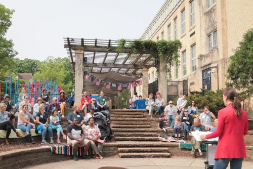 A group of people sitting on the steps in a small amphitheater while listening to someone speaking to the crowd.