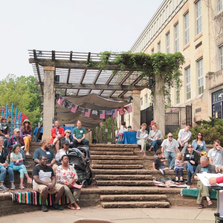 A group of people sitting on the steps in a small amphitheater while listening to someone speaking to the crowd.