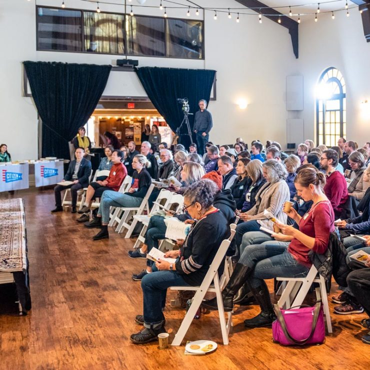 A large room with a crowd of people seated and listening to a person sharing from the stage.
