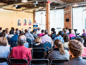A large room with a crowd of people seated and listening to a person sharing from the stage.