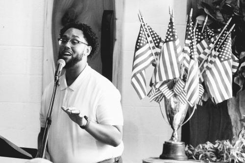 A person speaking into a microphone with American flags in the background.