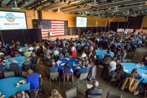 A large conference room, full of round tables with people sitting around them, as well as a stage with a podium and large American flag.