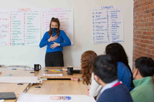 A person standing in front of a group of people at a table with their hands to their heart.