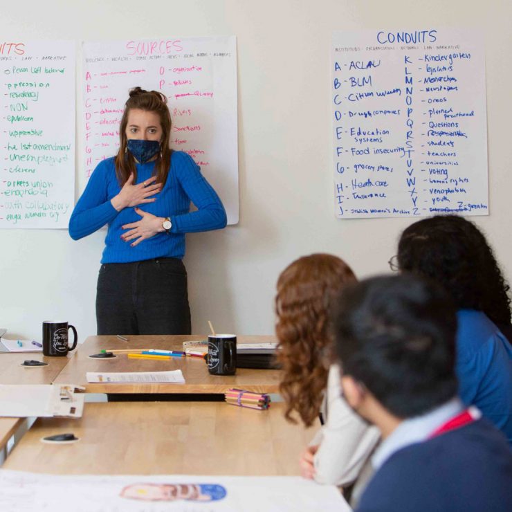 A person standing in front of a group of people at a table with their hands to their heart.