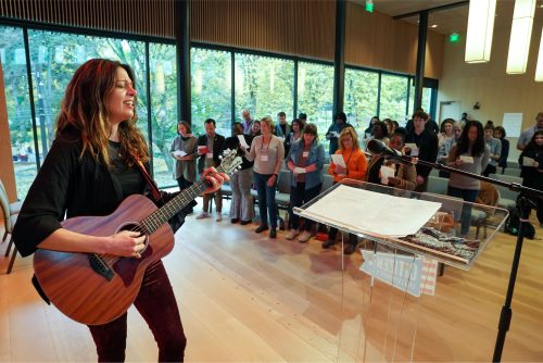 A large group standing and singing along with a person playing the guitar in the front of the room.