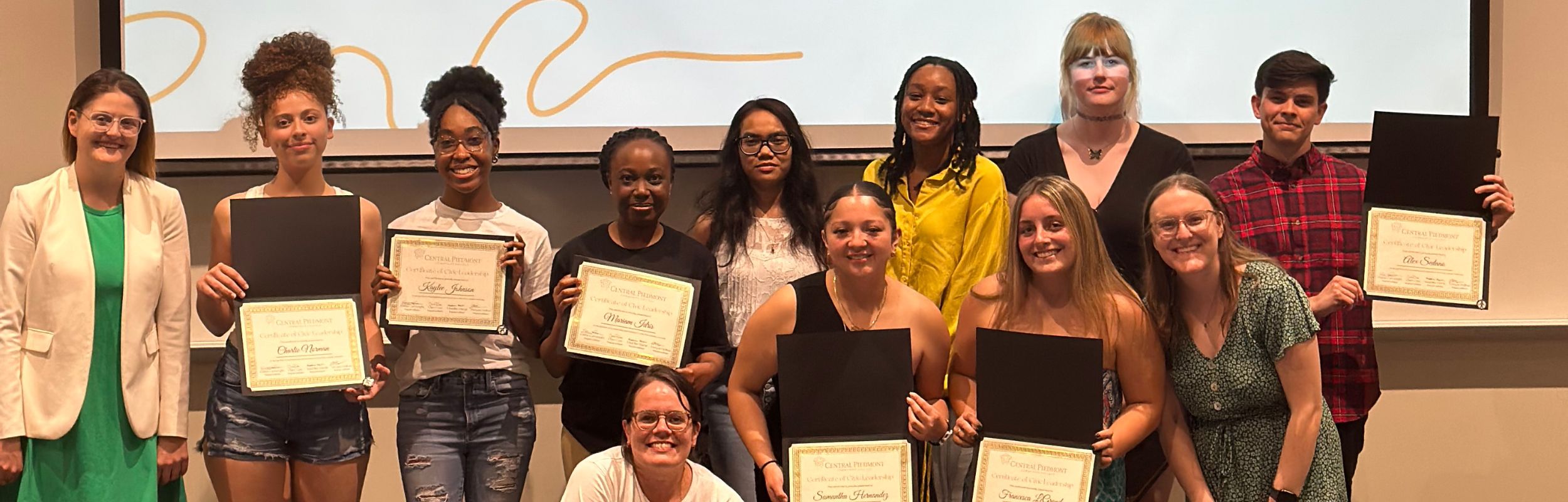 A group of students at the front of a room smiling and holding diplomas. 